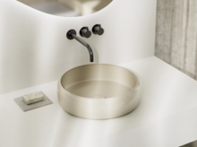Modernes, rundes Aufsatzwaschbecken in hellem Beige auf weißer Konsole mit schwarzer Wandarmatur und Seifenablage.