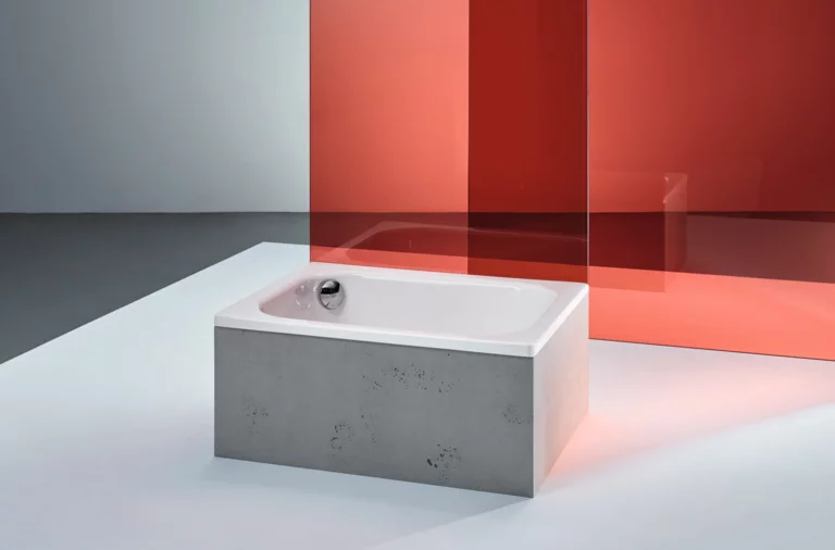 Rectangular steel-enamel bathtub with straight edges, freestanding in front of red glass panel on light floor.