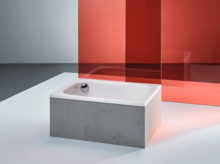 Rectangular steel-enamel bathtub with straight edges, freestanding in front of red glass panel on light floor.