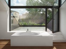 Rectangular steel-enamel bathtub with oval interior, freestanding in front of large windows, modern setting.