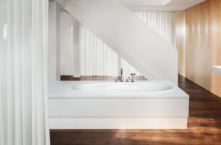 White rectangular steel-enamel bathtub with oval interior, in front of stairs, on dark wooden floor.
