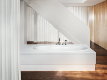 White rectangular steel-enamel bathtub with oval interior, in front of stairs, on dark wooden floor.