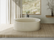 Freestanding oval bathtub in light beige on wooden platform, in front of window with curtain and minimal decor.