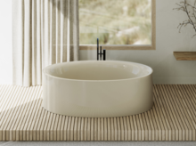 Freestanding oval bathtub in light beige on wooden platform, in front of window with curtain and minimal decor.
