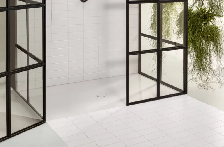 Superficie de ducha rectangular blanca de acero esmaltado con desagüe central, rodeada de paredes de vidrio, azulejos blancos.