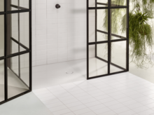 Superficie de ducha rectangular blanca de acero esmaltado con desagüe central, rodeada de paredes de vidrio, azulejos blancos.