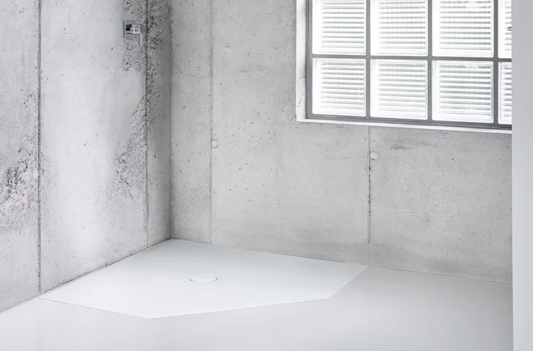 White, pentagonal steel enamel shower tray in modern, minimalist bathroom with concrete walls and large window.