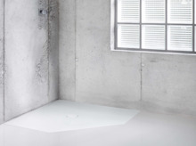 White, pentagonal steel enamel shower tray in modern, minimalist bathroom with concrete walls and large window.