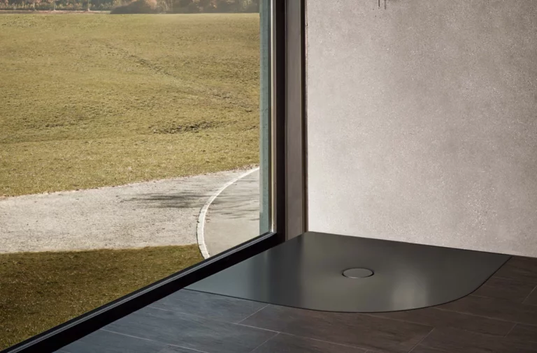 Superficie de ducha de acero vitrificado en color oscuro, forma redondeada y angular, a nivel del suelo frente a ventana y pared.