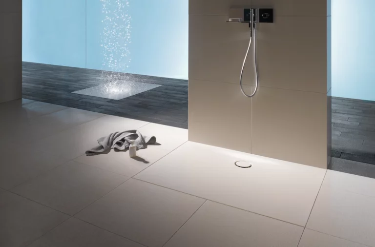 Прямоугольный стальной эмалированный душевой поддон вровень с полом, светлый, с настенным смесителем, рядом полотенце на плиточном полу.