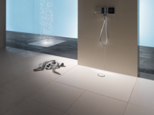 Прямоугольный стальной эмалированный душевой поддон вровень с полом, светлый, с настенным смесителем, рядом полотенце на плиточном полу.