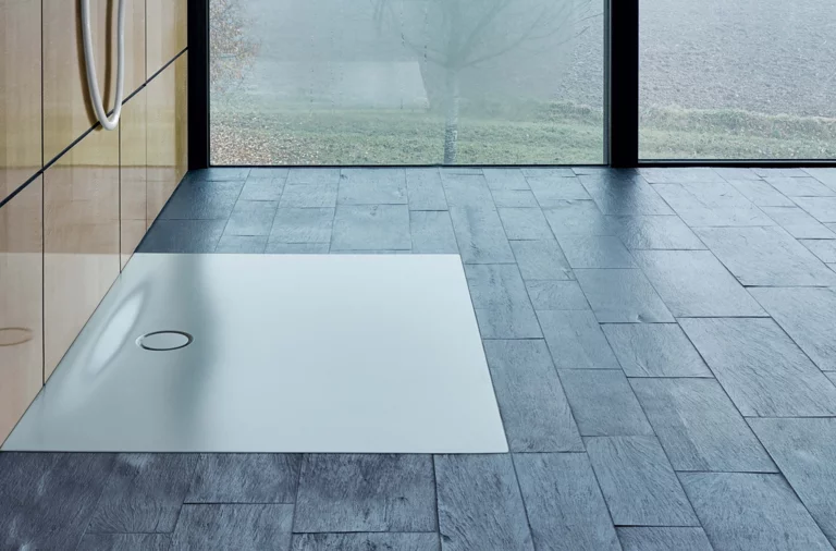 Прямоугольный, плоский душевой поддон из стальной эмали с центральным сливом, установлен вровень с полом на тёмной плитке.