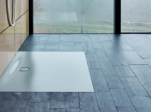 Прямоугольный, плоский душевой поддон из стальной эмали с центральным сливом, установлен вровень с полом на тёмной плитке.