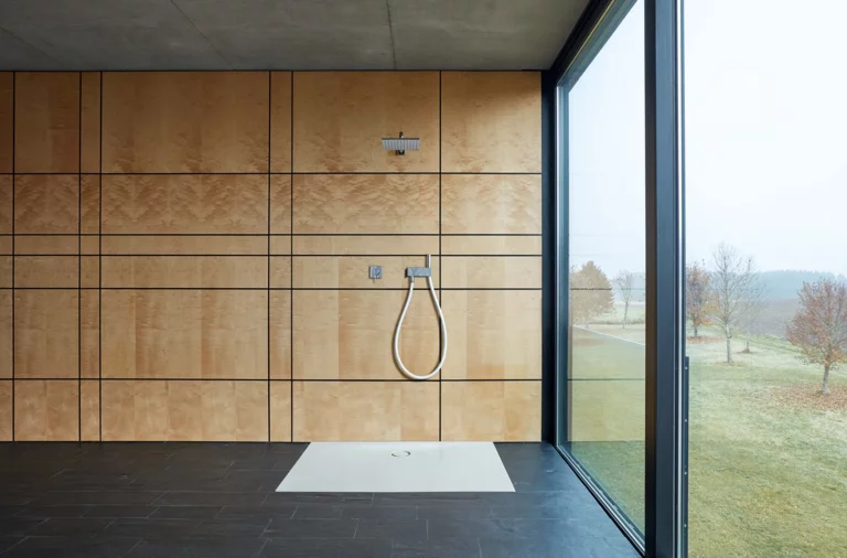 Прямоугольный душевой поддон из стальной эмали, вровень с полом, с ручным душем в минималистичной ванной с большим окном.