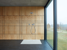 Прямоугольный душевой поддон из стальной эмали, вровень с полом, с ручным душем в минималистичной ванной с большим окном.