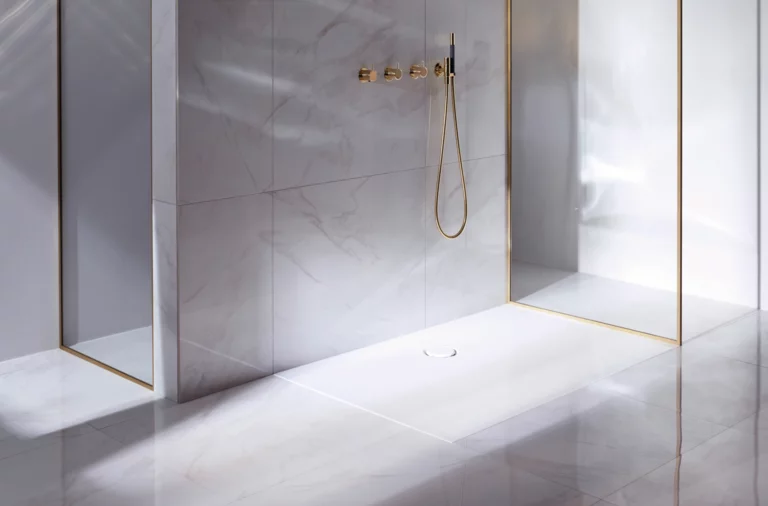 Прямоугольный душевой поддон из стальной эмали, вровень с полом, с золотистой арматурой и стеклянной перегородкой, окружён светлой плиткой.