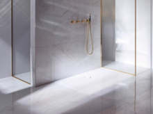 Прямоугольный душевой поддон из стальной эмали, вровень с полом, с золотистой арматурой и стеклянной перегородкой, окружён светлой плиткой.