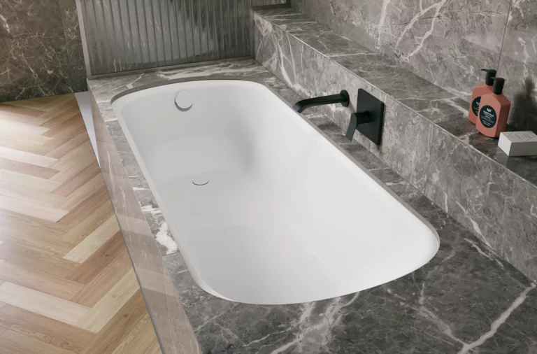 Bañera blanca de acero titanio esmaltado en entorno gris efecto piedra, con grifo mural negro.