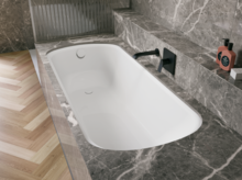 Bañera blanca de acero titanio esmaltado en entorno gris efecto piedra, con grifo mural negro.