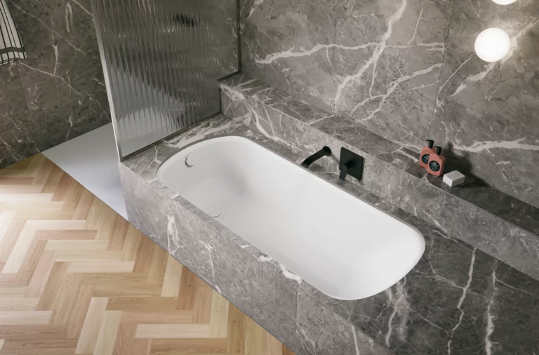 Bañera blanca de acero titanio esmaltado en entorno gris efecto piedra, con grifo negro y suelo en espiga.