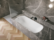 Bañera blanca de acero titanio esmaltado en entorno gris efecto piedra, con grifo negro y suelo en espiga.