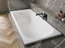 Bañera blanca de acero titanio esmaltado en entorno gris efecto piedra, con grifo mural negro.