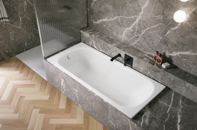 Bañera blanca de acero titanio esmaltado en entorno gris efecto piedra, con grifo negro y suelo en espiga.