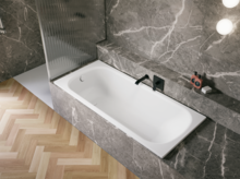 Bañera blanca de acero titanio esmaltado en entorno gris efecto piedra, con grifo negro y suelo en espiga.