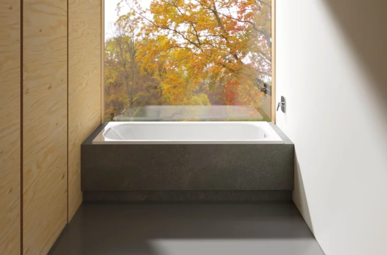 Rechteckige Badewanne aus Stahl-Email, in dunklem Podest vor großem Fenster mit Blick auf herbstliche Bäume.
