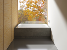 Rechteckige Badewanne aus Stahl-Email, in dunklem Podest vor großem Fenster mit Blick auf herbstliche Bäume.