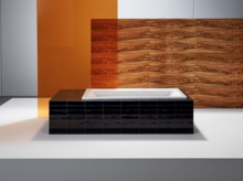 Rechteckige Badewanne aus Stahl-Email, eingefasst in schwarze Fliesen, vor Wand mit Holzoptik und orangefarbenem Glaspanel.