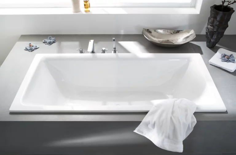 Rechthoekig bad van staal-email, ingebouwd in grijs oppervlak met kranen en witte handdoek op de rand.