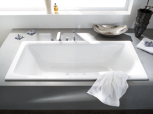 Rechthoekig bad van staal-email, ingebouwd in grijs oppervlak met kranen en witte handdoek op de rand.