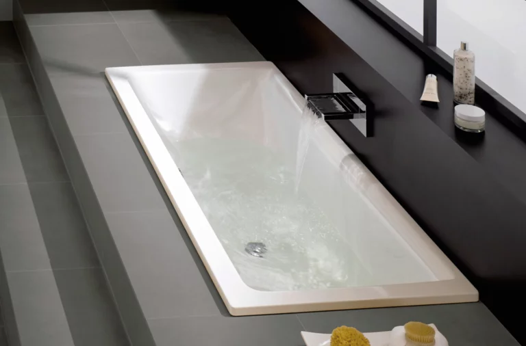 Rechteckige Badewanne aus Stahl-Email, eingelassen in graue Fliesen, wird mit Wasser aus einer Wandarmatur befüllt.