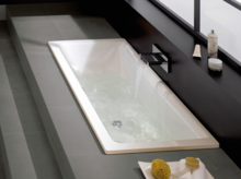 Rechthoekig bad van staal-email, verzonken in grijze tegels, wordt gevuld met water uit een wandkraan.