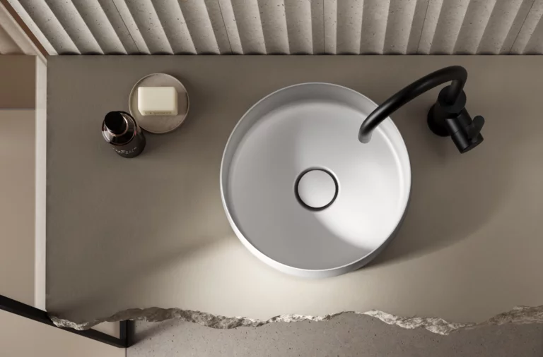 Ronde stalen emaille wastafel op inbouwblad met zwarte kraan, daarnaast zeep en fles, minimalistisch ontwerp.