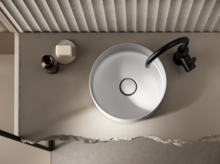 Ronde stalen emaille wastafel op inbouwblad met zwarte kraan, daarnaast zeep en fles, minimalistisch ontwerp.