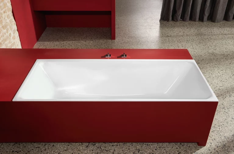 Rechteckige Badewanne aus Stahl-Email, eingebaut in roten Rahmen, vor Steinwand und Vorhang, auf grauem Boden.