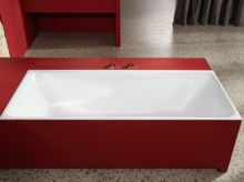 Rechteckige Badewanne aus Stahl-Email, eingebaut in roten Rahmen, vor Steinwand und Vorhang, auf grauem Boden.