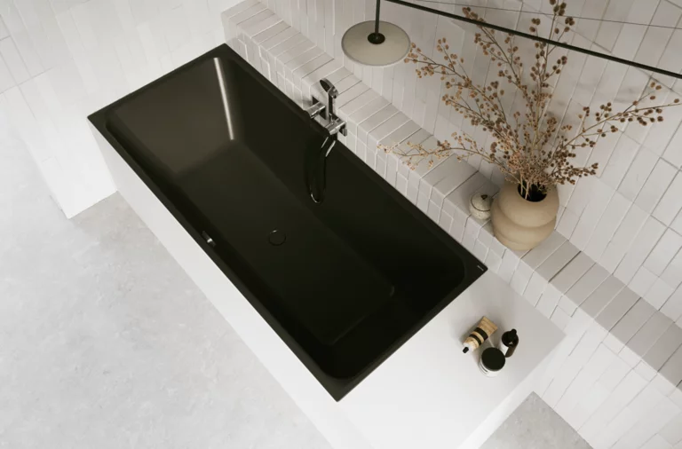 Schwarze Badewanne in hellem Bad mit weißen Fliesen, Armatur, Handtuch und Vase mit Zweigen.