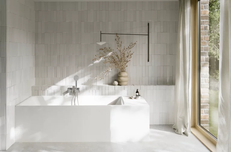 Weiße Badewanne in hellem Bad mit weißen Fliesen, Handbrause, Vase mit Zweigen und großem Fenster.