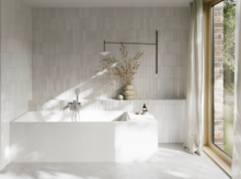 Weiße Badewanne in hellem Bad mit weißen Fliesen, Handbrause, Vase mit Zweigen und großem Fenster.