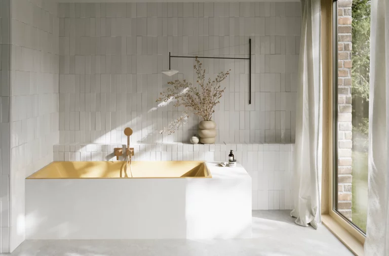 Goldfarbene Badewanne in hellem Bad mit weißen Fliesen, Handbrause, Vase mit Zweigen und großem Fenster.