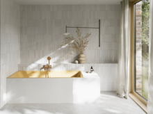 Goldfarbene Badewanne in hellem Bad mit weißen Fliesen, Handbrause, Vase mit Zweigen und großem Fenster.
