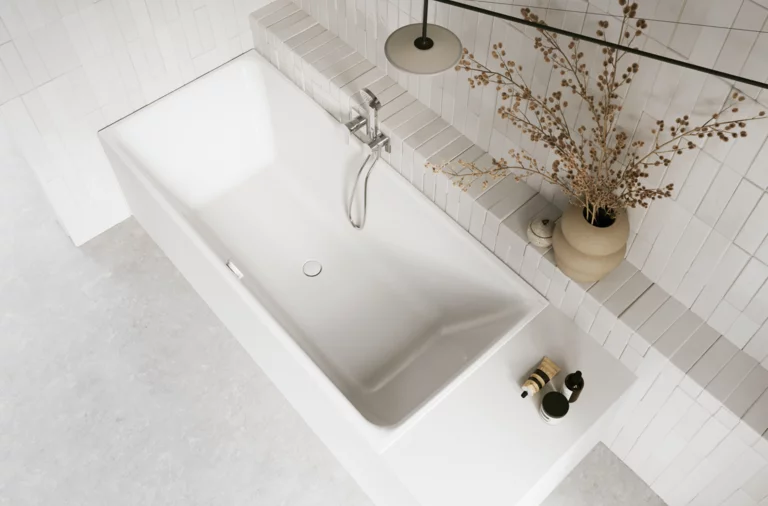 Weiße Badewanne in hellem Bad mit weißen Fliesen, Armatur, Handtuch und Vase mit Zweigen.