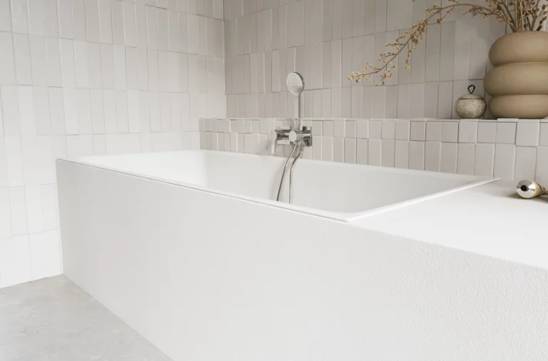 Weiße Badewanne in hellem Bad mit weißen Fliesen, Armatur, Handbrause und Vase mit Zweigen.