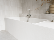 Weiße Badewanne in hellem Bad mit weißen Fliesen, Armatur, Handbrause und Vase mit Zweigen.