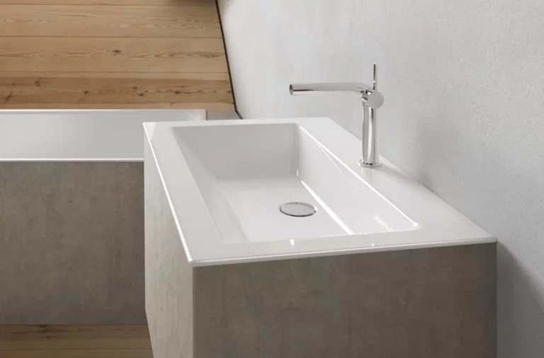 Rechteckiges Stahl-Email-Waschbecken auf Einbauplatte mit moderner Armatur, vor heller Wand und Holzfußboden.