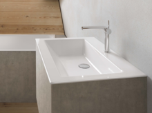 Rechteckiges Stahl-Email-Waschbecken auf Einbauplatte mit moderner Armatur, vor heller Wand und Holzfußboden.