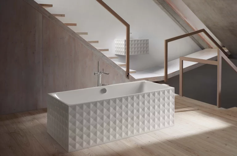 Rechteckige Badewanne aus Stahl-Email mit geometrischer Musterstruktur, freistehend auf Holzfußboden vor Treppe.